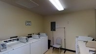 This single-floor laundry facility is designed with practicality and functionality at its core. The room features a row of top-loading washing machines and dryers neatly arranged along one wall, providing ample capacity for multiple users. Overhead, a long fluorescent light fixture ensures bright, even illumination throughout the space, making it easy to sort and manage laundry at any time of day. The walls are finished in a neutral cream tone, contributing to a clean and utilitarian atmosphere. A freestanding garment rack is positioned toward the center of the room, offering a convenient space to hang freshly laundered clothing. A folding chair nearby provides a spot to wait or attend to laundry tasks. An electrical panel is mounted on the wall, and a small waste bin is available for convenience. A posted housekeeping notice on the wall reflects an emphasis on maintaining cleanliness and shared responsibility within the space. The light-colored tile flooring and textured ceiling complete the no-frills, practical design of this communal laundry area, making it a straightforward and well-equipped space for residents or occupants to manage their laundry needs.