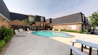 The Oaks offers a thoughtfully designed single-floor living space that blends artistic character with understated elegance. The interior is defined by polished concrete floors that lend an industrial-chic quality to the space, complemented by clean white walls that serve as a gallery-like backdrop for curated artwork displayed throughout. Recessed lighting provides warm, focused illumination across each room, enhancing the sophisticated ambiance. A standout feature of the dining area is a striking round green marble table set on a sculptural pedestal base, surrounded by slender rattan chairs and anchored by a woven area rug. A large-scale framed artwork adorns the wall nearby, reinforcing the space's artistic sensibility. Rustic, antique-style glass panel doors connect the rooms, adding a layer of vintage charm and visual depth. The interiors flow naturally between spaces, with additional wall-mounted artwork and framed pieces visible throughout, creating a cohesive gallery feel. Outside, residents have access to a generously sized outdoor swimming pool set within a well-maintained courtyard, enclosed by classic brick buildings with dark pitched roofs. Lounge chairs and patio seating surround the pool area, providing an inviting retreat. The overall atmosphere of The Oaks is one of refined, artistic living with comfortable community amenities.