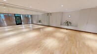 Located on Tresser Boulevard in Stamford, Connecticut, this fitness and movement studio offers a generously sized, open-plan space designed to accommodate a wide range of exercise and wellness activities. The studio is defined by its polished hardwood flooring, which runs the full length of the room and provides an ideal surface for yoga, dance, fitness classes, and more.

A standout feature of the space is its striking ombre accent wall, transitioning seamlessly from warm terracotta tones at the top to cool ocean blues at the base, adding a vibrant and calming visual element to the environment. Opposite this wall, floor-to-ceiling mirrors extend across the full width of the room, visually expanding the space and providing essential functional reflection for movement-based activities.

Recessed lighting throughout the ceiling ensures bright, even illumination across the entire studio. The entry side of the room is lined with elegant sheer curtains that soften the transition from the exterior glass partition, allowing natural light to filter in while maintaining a sense of privacy. Built-in white cabinetry along one wall provides practical storage, and subtle greenery adds a refreshing, natural touch. The overall ambiance is clean, modern, and welcoming — purpose-built for focused movement and wellness practice.