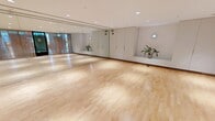 Located on Tresser Boulevard in Stamford, Connecticut, this fitness and movement studio offers a generously sized, open-plan space designed to accommodate a wide range of exercise and wellness activities. The studio is defined by its polished hardwood flooring, which runs the full length of the room and provides an ideal surface for yoga, dance, fitness classes, and more.

A standout feature of the space is its striking ombre accent wall, transitioning seamlessly from warm terracotta tones at the top to cool ocean blues at the base, adding a vibrant and calming visual element to the environment. Opposite this wall, floor-to-ceiling mirrors extend across the full width of the room, visually expanding the space and providing essential functional reflection for movement-based activities.

Recessed lighting throughout the ceiling ensures bright, even illumination across the entire studio. The entry side of the room is lined with elegant sheer curtains that soften the transition from the exterior glass partition, allowing natural light to filter in while maintaining a sense of privacy. Built-in white cabinetry along one wall provides practical storage, and subtle greenery adds a refreshing, natural touch. The overall ambiance is clean, modern, and welcoming — purpose-built for focused movement and wellness practice.