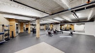 This substantial fitness facility spans a single floor and offers a generously proportioned exercise space designed to accommodate a wide range of workout activities. The interior carries a distinctive industrial aesthetic, characterized by exposed concrete ceilings with visible overhead pipework, structural columns clad in natural wood, and durable rubber flooring throughout that provides both safety and comfort underfoot.

The main exercise area is organized into several functional zones. A dedicated cardio section houses treadmills, stationary bikes, stair climbers, and rowing machines, while a strength training area features a comprehensive dumbbell rack, a Synrgy360 functional training system, weight benches, and kettlebells. A separate open floor zone with yoga mats and benches provides ample space for stretching, floor work, or group exercise. Large wall mirrors are strategically placed to allow users to monitor their form during workouts.

Additional storage for medicine balls and accessories is neatly arranged along the walls. The space is illuminated by bright overhead fluorescent strip lighting, ensuring clear visibility across all areas. Concrete masonry walls and the raw, unfinished ceiling contribute to the utilitarian yet purposeful atmosphere of this well-equipped fitness environment.