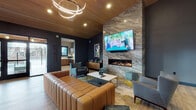 Located at 2930 Kinney Loop in Eugene, Oregon, this single-floor community clubhouse delivers a sophisticated and inviting atmosphere through a thoughtfully curated modern design. The main lounge area serves as the heart of the space, anchored by a dramatic floor-to-ceiling stone accent wall that houses both a linear gas fireplace and a wall-mounted flat-screen TV. Elevated ceilings clad in warm wood paneling add an organic, upscale touch, while a striking multi-ring contemporary chandelier provides ambient lighting overhead. Rich cognac leather sectional seating, plush accent chairs, and curated décor pieces contribute to a refined yet comfortable ambiance throughout. Wide-plank light hardwood flooring flows seamlessly across the entire space, complementing the deep charcoal-toned walls. Large black-framed windows invite ample natural light and frame views of the surrounding community. A dedicated game and social zone features a shuffleboard table alongside bistro-style seating arrangements, ideal for casual gatherings. The space also includes a well-appointed kitchenette outfitted with warm wood cabinetry, light countertops, a decorative tile backsplash, stainless steel appliances, and a utility sink. A welcoming entry area with tufted leather chairs and contemporary artwork completes this well-designed communal retreat.