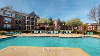 Located at 3701 Grapevine Mills Pkwy in Grapevine, Texas, this amenity space offers residents a well-appointed fitness center and a resort-style outdoor pool area. The fitness center is a visually striking space defined by its bold, multi-colored tile flooring in orange, teal, gray, and white geometric patterns, complemented by a coffered ceiling with vibrant orange accent panels and exposed gray beams. Ceiling fans and ample lighting throughout the room create a comfortable workout environment. The facility is equipped with a comprehensive selection of exercise machines, including Matrix treadmills, cable and weight machines, a stair climber, and a full dumbbell rack, alongside free weights and exercise balls. Full-wall mirrors visually expand the space, and multiple mounted flat-screen TVs provide entertainment during workouts. French doors and windows allow natural light to fill the room. The outdoor pool area features a generously sized, crystal-clear swimming pool with a surrounding deck appointed with teal lounge chairs and patio tables. Mature flowering trees and lush landscaping frame the space, while the backdrop of classic brick multi-story residential buildings adds a sense of community and architectural charm under the open Texas sky.