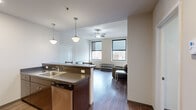 Located at 112 W Commerce St in downtown Youngstown, Ohio, this three-bedroom, two-bathroom unit offers a practical and modern layout across a single floor. The open-concept kitchen and living area serves as the heart of the space, where a peninsula with a dark granite-style countertop and bar seating connects the kitchen to the living area. The kitchen is equipped with stainless steel appliances, including a refrigerator, range, and microwave, complemented by rich dark cabinetry and recessed lighting. Pendant lights suspended over the peninsula add a contemporary touch. Hardwood-style flooring runs throughout the main living area, while a ceiling fan with light fixture in the living room enhances comfort. Large windows in the living area and bedrooms allow natural light to fill the space while offering views of the Youngstown urban skyline. The bedrooms feature neutral carpet flooring and additional windows with city views. Both bathrooms are finished with tile flooring, vanity cabinetry, and a tub-shower combination. The unit also includes a closet for additional storage. Clean, neutral walls throughout give the space a bright and versatile ambiance well-suited for urban living.