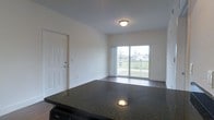 This single-floor space offers a clean, open-concept layout designed for comfortable everyday living. The living and kitchen areas flow seamlessly together, anchored by a striking dark granite countertop that serves as both a functional workspace and a visual centerpiece. The kitchen is equipped with standard appliances, adding practical convenience to the space.

The living area features hardwood-style flooring that extends throughout, providing a warm and cohesive aesthetic. Neutral white walls and a flush ceiling light fixture keep the interior bright and understated, allowing the architectural details to speak for themselves.

A standout feature of the space is the large sliding glass door that floods the interior with natural light and opens directly onto a private balcony. The balcony overlooks a green outdoor area, offering a pleasant outdoor retreat and scenic views of the surrounding landscape.

A solid entry door with a deadbolt lock provides a secure and clearly defined entrance to the unit. The overall design is straightforward and functional, with a light, airy ambiance created by the abundant natural light, clean lines, and open layout. The space presents a well-maintained interior ready for immediate use.