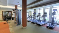 This modern fitness center is designed to support a comprehensive workout experience within a single, open-plan floor. The space is anchored by a row of treadmills positioned along a wall of large floor-to-ceiling windows, allowing natural light to pour in and providing views of the surrounding greenery — an energizing backdrop for cardio sessions. The light-toned flooring and neutral ceiling tones create a clean, functional aesthetic throughout. Structural columns with warm terracotta-colored accents add an architectural detail that breaks up the open layout without interrupting the flow of the space. On the strength training side, a multi-station cable machine and additional resistance equipment offer a variety of workout options, complementing the cardio zone effectively. A rowing machine is also visible, rounding out the range of available fitness equipment. Recessed lighting supplements the abundant natural light, ensuring the space remains bright and well-lit at all times. The overall ambiance is professional and inviting, with a practical layout that provides ample room to move between equipment. This fitness center is well-suited for users seeking a fully equipped facility for both cardiovascular and strength-based training.