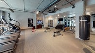 Located at 1041 Jo Johnston Ave in Nashville's vibrant 37203 corridor, this fitness center offers a thoroughly outfitted workout environment across a single floor. The main exercise area features an industrial-inspired aesthetic, with exposed concrete ceilings, visible ductwork, and neutral-toned carpet flooring that lends a modern, urban feel to the space.

The cardio zone is lined with a row of treadmills and elliptical machines, each equipped with individual screens for entertainment. Large windows along this section of the facility bring in abundant natural light and offer views of the surrounding neighborhood, creating an energizing atmosphere for workouts.

The strength training area is stocked with a full dumbbell rack, multiple weight benches, a cable machine system, and a functional training rig with resistance bands and accessories. Mirrored walls throughout this zone enhance the sense of space and allow for form monitoring during exercise. A mounted flat-screen television and a beverage refrigeration unit are also present, adding convenience to the workout experience.

A separate studio room, accessed through large sliding barn-style doors with frosted glass panels, provides a dedicated space for cycling bikes and rowing machines. This room features smooth flooring and a ceiling fan, making it well-suited for high-intensity or group-style training sessions. The overall layout is thoughtfully organized to support a wide range of fitness activities.