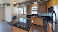 This single-floor unit offers a well-designed open-concept layout that seamlessly connects the living area to the kitchen. The spacious main living area features wood-look vinyl flooring, neutral white walls, and exposed industrial-style ductwork running along the ceiling, lending the space a contemporary urban character. Multiple windows allow generous natural light to fill the room, with views of the surrounding cityscape.

The kitchen is a standout feature, outfitted with warm maple cabinetry, striking dark granite countertops, and a large central island that provides additional prep and serving space. Stainless steel appliances — including a gas range, built-in microwave, and dishwasher — complete the functional and stylish cooking space. Wood-look flooring continues throughout the kitchen, maintaining a cohesive aesthetic.

The bedroom is carpeted for comfort, featuring clean white walls and a bright window fitted with blinds that offers views of the urban streetscape below. The full bathroom includes a tub-shower combination, a wood-toned vanity with a white countertop, a large mirror, and wood-look flooring, providing a clean and practical finish. Overall, the space blends industrial design elements with comfortable, modern living essentials.