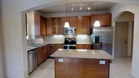 This thoughtfully designed space spans three floors, offering generous room to live and move comfortably. The kitchen serves as a standout feature, showcasing warm medium-toned wood cabinetry that extends to the ceiling, providing ample storage. Granite countertops in soft earth tones complement the rich cabinetry and extend to a central island, which offers additional workspace and built-in outlets for convenience. Stainless steel appliances — including a French door refrigerator with water and ice dispenser, a stainless range with electric cooktop, a built-in microwave, and a dishwasher — lend a sleek, modern feel to the culinary space. A tasteful diagonal tile backsplash adds visual interest behind the range, while pendant lighting over the island and a track lighting system overhead ensure the kitchen is well-lit and functional. Light tile flooring runs throughout, brightening the space and creating a clean, cohesive aesthetic. Glass-front upper cabinets near the sink add an elegant display element. The overall ambiance of the kitchen is warm yet contemporary, balancing practicality with refined design details across all three levels of the property.