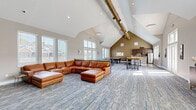 This single-floor community clubhouse offers a generously sized, open-concept layout designed for gathering and socializing. The space is defined by striking vaulted ceilings that rise dramatically overhead, creating an airy, expansive atmosphere throughout the main area. Large windows line the walls, flooding the interior with natural light and providing views of the surrounding neighborhood.

The main lounge area serves as the centerpiece of the space, featuring a large sectional leather sofa positioned in front of a wall-mounted flat-screen TV set against a bold teal accent wall — a vibrant design detail that adds personality to the room. Patterned carpet tile flooring flows throughout, complementing the neutral gray and warm beige tones of the walls.

A dedicated dining and bar area offers high-top tables with rustic live-edge surfaces alongside bar stools, providing casual seating for socializing or dining. Adjacent to this is a well-appointed kitchenette featuring dark espresso cabinetry, white quartz countertops, a stainless steel sink, dishwasher, and a coffee station. A bar-height counter with vibrant yellow upholstered stools connects the kitchen to the main area. The space is further enhanced by curated artwork and decorative accents, lending a welcoming, well-designed ambiance throughout.