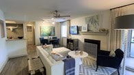 This single-floor space showcases an open-concept layout that seamlessly integrates the living and dining areas into one cohesive environment. The living area is anchored by a classic fireplace with a painted mantel, above which a flat-screen television is mounted, flanked by decorative artwork and accent pieces. A ceiling fan with integrated lighting occupies the center of the room, promoting airflow and comfort. The space is furnished with a tufted white sofa accented by colorful throw pillows, a round white coffee table, and a navy accent chair, all arranged atop a bold chevron-patterned area rug. Track lighting along the ceiling provides additional illumination. The dining area, positioned adjacent to the living space, features a compact table surrounded by navy upholstered chairs beneath a landscape painting that adds a vibrant pop of color. A window dressed with sheer curtains allows natural light to filter in. Dark hardwood-style flooring runs throughout, providing a warm contrast to the neutral wall tones. The overall ambiance is contemporary and well-appointed, balancing comfort with a cohesive, stylish aesthetic.