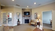 Located at 103 Melville Loop in Chapel Hill, NC, this single-floor space offers a welcoming and well-appointed layout designed for both comfort and functionality. The main living area serves as the centerpiece of the space, showcasing a striking floor-to-ceiling stacked stone fireplace with a wood mantel and wall-mounted television above. Crown molding runs throughout, lending a refined architectural detail to the warm, neutral-toned interiors. Plush carpeting in the lounge areas transitions to hardwood flooring near the entry foyer, where elegant French doors with glass panes open to a raised platform area filled with natural light from large windows. The kitchen is thoughtfully designed with warm wood cabinetry, stone tile backsplash, stainless steel appliances including a dishwasher and refrigerator, light tile flooring, and pendant lighting over a pass-through counter. Adjacent to the kitchen, a bright dining nook features a round table surrounded by woven chairs and oversized windows overlooking lush greenery outdoors. A private office or meeting room provides a quieter setting with carpeted floors and crown molding. Throughout the space, recessed lighting, large windows, and a cohesive warm color palette create an inviting and functional atmosphere suited for a variety of uses.