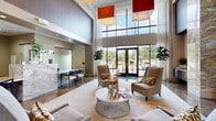 This elegantly designed single-floor amenity space makes an immediate impression with its dramatic double-height entry lobby, where soaring ceilings are accentuated by bold, oversized pendant light fixtures in striking red and orange hues. The polished herringbone-pattern stone flooring reflects abundant natural light pouring through floor-to-ceiling glass entry doors and expansive clerestory windows, creating a bright and welcoming atmosphere.

The main lobby area features a refined seating arrangement with neutral-toned upholstered wingback chairs centered around a sculptural round coffee table, ideal for relaxed conversation. A sleek marble-topped reception desk anchors one side of the lobby, framed by frosted glass partitions that lend a sense of privacy while maintaining visual openness.

Adjacent lounge areas offer additional gathering spots, each distinctly styled. One features contemporary metallic accent chairs positioned beneath a wall-mounted flat-screen television, set against warm wood-paneled accents. Another area showcases a substantial marble-topped bar island illuminated by a row of elegant cylindrical pendant lights, with dark cabinetry and a built-in media console as a backdrop.

A separate dining nook with large windows overlooking the outdoors provides a bright, community-oriented workspace or gathering area, complete with a long communal table, woven leather barstools, and a modern orbital chandelier. Throughout, lush greenery and curated décor add warmth and sophistication to this polished, contemporary communal space.