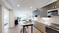 This single-floor residence showcases a thoughtfully designed open-concept layout connecting the kitchen, dining, and living areas in one cohesive space. The kitchen is outfitted with two-tone cabinetry—combining dark wood upper cabinets with lighter lower units—complemented by a white subway tile backsplash, white quartz countertops, and a full suite of stainless steel appliances including a built-in microwave, electric range, dishwasher, and refrigerator. Light wood-look flooring runs throughout the main living area, enhancing the bright, airy atmosphere created by recessed lighting overhead. A bar-height dining table positioned between the kitchen and living area provides a casual space for dining or entertaining.

The bedroom is a serene retreat finished with plush carpeting and neutral tones. Sliding glass doors open directly onto a private balcony, allowing abundant natural light to fill the room. The full bathroom features a wood-grain vanity with a white countertop, a large mirror, built-in shelving, and a soaking tub with shower combination. Clean lines, neutral palettes, and modern fixtures are consistent throughout, creating a cohesive and contemporary aesthetic across the entire space.