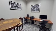 This single-floor business center is designed to provide a focused and functional environment for work and productivity. The space features a long wooden wall-mounted desk that comfortably accommodates two workstations, each equipped with a desktop computer — including both a PC and an Apple iMac — catering to a range of user preferences. A multifunction printer sits conveniently between the two stations, offering printing, scanning, and copying capabilities. Patterned carpeting in cool grey and blue tones adds a professional yet approachable aesthetic to the room. A round wooden high-top table with bar-height chairs is positioned to the side, offering an alternative informal seating area for quick meetings or casual work. The walls are finished in a neutral light grey and are adorned with two large black-and-white framed photographs — one depicting a classic steering wheel and the other a striking urban architectural scene — lending the space a refined, contemporary character. The overall ambiance is clean, organized, and professional, making this business center a practical and inviting resource for those seeking a dedicated workspace.