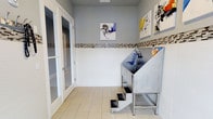 Located at 1871 Ashley River Road in Charleston, South Carolina, this single-floor pet grooming facility is thoughtfully designed to provide a clean, professional, and welcoming environment for pets and their owners. The space features a dedicated grooming area equipped with a stainless steel dog bathing tub complete with built-in entry steps, making it accessible and comfortable for pets of all sizes. A coiled utility hose and grooming supplies are conveniently within reach, reflecting the functional layout of the workspace. The walls are finished with crisp white tile on the lower half, accented by a decorative mosaic tile border that adds a refined touch to the utilitarian space. The upper walls are painted in a soft gray tone, lending a calm and neutral ambiance throughout. Light-colored ceramic tile flooring runs underfoot, contributing to the clean and hygienic character of the facility. Animal-themed artwork adorns the walls, including expressive black-and-white illustrations and a colorful mixed-media piece, adding personality and charm to the environment. A glass-paneled door provides visibility between areas, enhancing the open feel of the space. Overall, the facility strikes a balance between professional functionality and an inviting aesthetic.