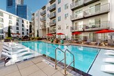 This contemporary urban residential community is designed around a thoughtfully curated outdoor amenity deck that serves as the heart of the property. The centerpiece is a generously sized resort-style swimming pool, framed by a wide sun-drenched deck lined with lounge chairs and bold red market umbrellas, creating an inviting atmosphere for relaxation. The pool area transitions seamlessly into a landscaped courtyard featuring raised stone planting beds bursting with colorful seasonal flora, all shaded by a sleek steel pergola strung with overhead lighting. Adjacent to the pergola, a casual seating area with plush lounge chairs and low tables provides a comfortable gathering space. A dedicated lawn area offers room for outdoor games, complete with cornhole boards and Adirondack-style seating arranged around a central fire feature. The community also includes a semi-covered lobby-level lounge with soaring double-height ceilings, floor-to-ceiling glass walls, stone accent detailing, and an open flow to the outdoors. An elevated terrace off the leasing center provides additional alfresco seating with views of the surrounding urban skyline. Vibrant potted plantings and manicured landscaping accent the building's clean, modern exterior throughout.