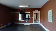 This single-floor fitness center is thoughtfully designed to support an effective workout experience. The space is defined by deep, rich red-toned walls that create an energizing atmosphere, complemented by white trim and a clean white ceiling. Durable dark rubber flooring covers the main workout area, providing both safety and functionality underfoot.

A large floor-to-ceiling mirror spans one wall, visually expanding the space and allowing users to monitor their form during exercise. Reflected in the mirror is a multi-function strength training cable machine, along with additional fitness equipment, making efficient use of the available area. Natural light filters in through the windows visible in the mirror's reflection, contributing to a bright and welcoming environment.

A wall-mounted flat-screen television is positioned prominently above the mirror, offering entertainment or instructional content during workouts. Overhead, a rectangular fluorescent light fixture provides consistent, even illumination throughout the space.

A white-framed glass-panel door leads to an adjacent room — visible through the door is a carpeted hallway or secondary space, suggesting convenient access to additional amenities. A wall-mounted HVAC vent ensures comfortable climate control year-round. Overall, this fitness center delivers a focused and well-equipped environment suitable for strength and general training.