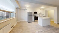 This 2-bedroom, 1-bathroom unit offers a thoughtfully laid-out single-floor plan with a clean, modern aesthetic throughout. Light-toned laminate flooring runs continuously across all rooms, contributing to a bright and cohesive feel. The open-concept living and kitchen area serves as the heart of the unit, where large, wide windows flood the space with natural light and provide outdoor views of surrounding greenery. The kitchen is well-appointed with white shaker-style cabinetry, stainless steel appliances including a refrigerator, electric range, and over-the-range microwave, a white countertop island, and a tile backsplash — offering both functionality and a modern look. The living area provides generous room for furnishings and flows seamlessly from the kitchen. Both bedrooms are comfortably sized, each featuring large windows with radiator heating units beneath them and sliding-door closets for storage. The primary bedroom is notably spacious. The shared bathroom is finished with clean white subway tile surrounding a full soaker tub, a white vanity, and chrome fixtures. A hallway connects the bedrooms and bathroom, completing this efficiently organized and move-in-ready layout.
