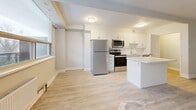This 2-bedroom, 1-bathroom unit offers a thoughtfully laid-out single-floor plan with a clean, modern aesthetic throughout. Light-toned laminate flooring runs continuously across all rooms, contributing to a bright and cohesive feel. The open-concept living and kitchen area serves as the heart of the unit, where large, wide windows flood the space with natural light and provide outdoor views of surrounding greenery. The kitchen is well-appointed with white shaker-style cabinetry, stainless steel appliances including a refrigerator, electric range, and over-the-range microwave, a white countertop island, and a tile backsplash — offering both functionality and a modern look. The living area provides generous room for furnishings and flows seamlessly from the kitchen. Both bedrooms are comfortably sized, each featuring large windows with radiator heating units beneath them and sliding-door closets for storage. The primary bedroom is notably spacious. The shared bathroom is finished with clean white subway tile surrounding a full soaker tub, a white vanity, and chrome fixtures. A hallway connects the bedrooms and bathroom, completing this efficiently organized and move-in-ready layout.