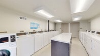 Located at 308 Scott St SW in Atlanta, GA, this shared laundry facility is a clean, efficiently designed single-room space that offers a comprehensive suite of laundry amenities. The room accommodates a generous row of top-load washing machines and front-load dryers, all white and uniformly arranged along the perimeter walls, providing ample capacity for high-volume use. Each machine is equipped with a digital payment interface for convenient, cashless operation. At the center of the room stands a prominent island with a marble-style countertop and dark navy base, offering a dedicated surface for sorting and folding laundry. The space is bathed in bright, even light from large rectangular flush-mount ceiling fixtures, ensuring excellent visibility throughout. Light-toned flooring and crisp white walls contribute to a clean, welcoming atmosphere. A large glass door on one end of the room floods the space with natural light and provides views of the surrounding greenery, while also serving as a secondary egress. Informational signage is posted on the walls for user guidance. The facility also features a visible electrical panel, ventilation grilles, and a fire safety system, reflecting attention to both functionality and safety standards.