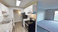 This single-floor residence offers a thoughtfully designed layout that balances functionality and comfort. The kitchen is a standout feature, showcasing crisp white shaker-style cabinetry that extends to the upper walls, providing ample storage space. Light granite-toned countertops complement the cabinetry, while the kitchen is equipped with a stainless steel refrigerator and a black and stainless electric range with a built-in overhead vent hood. A skylight-style flush ceiling fixture floods the kitchen with bright, even lighting, enhancing the clean and airy atmosphere. The flooring throughout the kitchen and adjoining areas consists of warm-toned wood-look planks, adding a modern yet inviting touch. The kitchen opens naturally toward the living area, which features plush carpeting in a neutral gray tone, creating a cozy and comfortable environment. A textured ceiling adds architectural character throughout the space. A hallway visible from the kitchen leads to additional rooms, suggesting a practical and well-connected floor plan. The overall ambiance is fresh, clean, and move-in ready, with updated finishes that give the space a contemporary residential feel.
