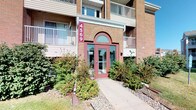 This residential building presents a classic brick facade accented by a distinctive arched entryway with a deep red door, framed by lush landscaping that adds inviting curb appeal. The building's exterior features multiple balconies and large windows, hinting at the comfortable living spaces within. The single-floor unit layout is designed for practical, everyday living. The bathroom is a standout feature, finished with light pink wall tiles surrounding a full soaking tub and shower combination equipped with both a fixed showerhead and a handheld spray. A large vanity mirror with globe-style Hollywood lighting illuminates the sink area, which is fitted with a single-basin countertop and warm wood-toned cabinetry. Neutral-toned tile flooring adds durability and a clean aesthetic throughout the bathroom. A notable convenience is the in-unit stacked washer and dryer, visible from the bathroom area, offering residents the ease of laundry without leaving home. The overall color palette throughout is soft and neutral, with white walls and dark wood trim creating a clean, understated aesthetic. The space is thoughtfully designed to balance functionality with comfort, making it a practical and welcoming residential unit.