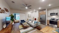 This thoughtfully designed single-floor residence offers a seamless blend of modern style and functional living. The open-concept main living area is anchored by warm hardwood-style flooring, a ceiling fan with integrated lighting, and recessed fixtures that ensure consistent illumination throughout. A sliding barn door connects the living space to the bedroom, adding a contemporary architectural detail. The living room accommodates a generous seating arrangement, a glass-top coffee table, and a dedicated media wall with a sleek wooden TV console, complemented by decorative wall art that enhances the room's character. A dining nook with a compact table and chairs sits adjacent to the living area, ideal for everyday meals. The kitchen features a dual-tone cabinetry design — warm wood lower cabinets paired with crisp white uppers — along with stainless steel appliances including a side-by-side refrigerator with ice and water dispenser, an electric range, microwave, and dishwasher, all centered around light quartz countertops. The bedroom is a serene retreat with large windows overlooking an outdoor terrace, allowing ample natural light and outdoor views. The bathroom is finished with wood-toned cabinetry, a single-basin vanity, a framed mirror, and abundant natural light from a sizeable window.