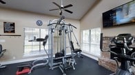 This dedicated fitness room offers a practical and comfortable environment for a full-body workout. The space is anchored by a multi-station cable strength machine, providing a wide range of resistance training options, and is complemented by cardio equipment including a treadmill. Rubber flooring throughout ensures safety and durability during exercise. Large windows fitted with horizontal blinds line the walls, allowing generous natural light to fill the room while offering views of the surrounding greenery outdoors. A ceiling fan overhead provides airflow and comfort during workouts, while a wall-mounted flat-screen television adds an entertainment option. A decorative brick accent wall adds a touch of warmth and texture to the otherwise neutral, light-toned interior. The vaulted ceiling contributes to an open, airy feel. A round analog clock on the wall keeps users mindful of their workout time. The overall design is functional and well-organized, creating an inviting atmosphere for both strength training and cardio exercise in a bright, well-ventilated setting.