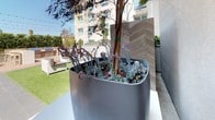 This thoughtfully designed outdoor amenity courtyard offers a resort-inspired retreat nestled within a contemporary multi-story residential building. The space is anchored by a sleek, low-profile concrete fire pit table surrounded by built-in bench seating accented with colorful throw pillows in blue, orange, and teal — creating an inviting central gathering area. Overhead, a canopy of bistro string lights adds warmth and ambiance, making the space equally enjoyable day and evening.

The courtyard is finished with large-format porcelain tile pavers that lend a clean, modern aesthetic, while a section of lush artificial turf provides a softer, casual zone. A dedicated outdoor kitchen and bar area features a built-in grill and high-top seating, ideal for entertaining. Dining tables and chairs are positioned nearby for al fresco meals.

Abundant greenery, including tall ornamental grasses, tropical palms, succulents, and flowering plants in oversized modern planters, lines the perimeter and introduces a vibrant, natural contrast to the neutral tones of the hardscape. Elevated planter beds with warm wood-toned cladding reinforce the sophisticated landscaping design. Elevator access is conveniently located at the courtyard's edge, ensuring seamless connectivity to the rest of the building.