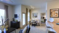 This single-story space offers an open and well-connected layout that seamlessly integrates the living, dining, and bedroom areas into one cohesive environment. The living area is anchored by a sleek leather sofa and a wall-mounted flat-screen television positioned atop a dark wood media console. Track lighting mounted on the ceiling provides focused illumination throughout the main living space, complementing the natural light that filters in through multiple windows dressed with green curtains and slatted blinds. The dining area features a dark wood table surrounded by upholstered chairs, with a decorative mirror serving as a focal point on the wall above. Neutral, light-toned carpeting extends throughout the space, contributing to a warm and inviting ambiance. A glimpse into the adjacent bedroom reveals a neatly arranged sleeping area with a green-accented bedspread, framed artwork, and a bedside lamp, suggesting a cohesive design aesthetic carried throughout. The warm beige walls and layered lighting create a comfortable and functional atmosphere suited to everyday living.