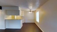 This single-floor space offers a practical and functional layout centered around an open-concept design that connects the kitchen and living area. The living space is anchored by rich, dark hardwood-style flooring that extends throughout, creating a warm and cohesive aesthetic. A ceiling fan with an integrated light fixture is centrally positioned overhead, providing both air circulation and ambient lighting to the room.

The kitchen features white cabinetry, a light-colored countertop, and a peninsula-style breakfast bar that serves as a natural divider between the cooking and living areas. The overhead range hood adds a structural focal point to the kitchen zone. White appliances complement the clean, bright cabinetry.

The living area benefits from a window fitted with blinds, allowing for adjustable natural light to fill the space. Neutral-toned walls paired with white trim and baseboards give the interior a clean, understated appearance. The textured popcorn ceiling adds a classic residential character throughout.

Overall, the space presents a straightforward, well-organized single-level layout that maximizes the connection between functional zones, making it an efficient and comfortable living environment.
