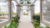 Located at 1456 Serfas Club Dr in Corona, California, this outdoor community amenity space offers a well-appointed pool area designed for relaxation and recreation. The centerpiece is a generously sized swimming pool with clear turquoise water, surrounded by a broad concrete deck furnished with teal lounge chairs and vibrant orange accent cushions and seating. A distinctive white pergola structure with textured stucco columns and an open gabled roof frames the entrance pathway, flanked by lush landscaping including tropical plants and ornamental greenery that enhance the resort-like atmosphere. Adjacent to the pool, an outdoor grilling station provides a convenient space for outdoor cooking and entertaining. The surrounding area is anchored by a Spanish Colonial-style clubhouse building, featuring a characteristic tile roof, white stucco exterior, and an iconic bell tower that adds architectural character to the setting. Tall pine trees provide natural shade and privacy, while the backdrop of rolling hills and open sky contributes to a scenic and tranquil environment. Multiple seating configurations, including poolside loungers and patio furniture with umbrellas, accommodate a range of outdoor activities throughout the day.
