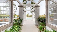 Located at 1456 Serfas Club Dr in Corona, California, this outdoor community amenity space offers a well-appointed pool area designed for relaxation and recreation. The centerpiece is a generously sized swimming pool with clear turquoise water, surrounded by a broad concrete deck furnished with teal lounge chairs and vibrant orange accent cushions and seating. A distinctive white pergola structure with textured stucco columns and an open gabled roof frames the entrance pathway, flanked by lush landscaping including tropical plants and ornamental greenery that enhance the resort-like atmosphere. Adjacent to the pool, an outdoor grilling station provides a convenient space for outdoor cooking and entertaining. The surrounding area is anchored by a Spanish Colonial-style clubhouse building, featuring a characteristic tile roof, white stucco exterior, and an iconic bell tower that adds architectural character to the setting. Tall pine trees provide natural shade and privacy, while the backdrop of rolling hills and open sky contributes to a scenic and tranquil environment. Multiple seating configurations, including poolside loungers and patio furniture with umbrellas, accommodate a range of outdoor activities throughout the day.