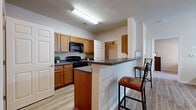 This single-floor residential unit offers a functional and well-organized layout across several distinct living areas. The kitchen is a standout feature, equipped with warm oak cabinetry, dark countertops, and black appliances including a range and over-the-range microwave. A peninsula-style breakfast bar with bar seating provides a casual dining option and creates a natural boundary between the kitchen and the rest of the living space. Light wood-style flooring flows throughout the main areas, lending a cohesive and clean aesthetic. The living room is generously sized, featuring vaulted ceilings, a ceiling fan, and large windows that allow abundant natural light and views of the surrounding outdoor environment. A secondary living or flex space offers additional room with its own wood-style flooring. The bedrooms are carpeted for comfort and each includes a ceiling fan and window, with one room configured to accommodate a desk workspace. The bathroom includes a full soaking tub and shower combination with clean white surround walls. A dedicated laundry room houses a full-size washer and dryer alongside wire shelving for storage, adding significant practical value. Overall, the unit presents a straightforward and comfortable residential environment.