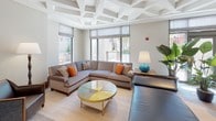 This single-floor space is defined by its striking coffered ceilings, which carry consistently throughout the main living and dining areas, lending architectural depth and a refined character to the interior. The layout is thoughtfully organized into distinct functional zones that flow naturally into one another.

The living area features a generously sized sectional sofa in a neutral gray upholstered fabric, accented with colorful throw pillows, centered around a gold-framed art glass coffee table. Large windows with Roman shades provide abundant natural light and views of the surrounding urban environment, while a secondary seating nook adjacent to the windows offers a more intimate retreat.

The dining area is anchored by a substantial dark-topped dining table surrounded by gold-framed upholstered chairs, illuminated by a cluster of globe pendant lights overhead. Adjacent to it, a sleek wet bar features a dark stone countertop, brass-finished banded detailing, and black-and-gold bar stools, adding a glamorous focal point.

The kitchen is equipped with a full suite of stainless steel Bosch appliances, including double wall ovens, a drawer microwave, and warm wood cabinetry with stone countertops. A separate lounge area provides additional seating alongside a console table with greenery and a blue ceramic accent lamp.