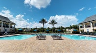 This outdoor pool area offers a well-appointed retreat designed for relaxation and recreation. The centerpiece is a generously sized swimming pool with clear, blue water, surrounded by an expansive paver deck finished in a warm, multi-toned brick pattern that lends a polished, resort-like aesthetic to the space.

Poolside, a collection of lounge chairs and blue shade umbrellas are arranged symmetrically, providing comfortable spots for sunbathing and leisure. Tall, mature palm trees frame the pool area, adding a tropical ambiance while complementing the open sky views above. The surrounding landscape includes lush greenery and views of the neighboring community buildings, contributing to a serene outdoor atmosphere.

Adjacent to the pool deck is a covered pavilion structure featuring arched stucco columns, ornamental ironwork fencing, and ceiling fans—offering a shaded gathering space for socializing or dining. The pavilion connects seamlessly to the pool area through a secured iron gate entry, reinforcing both the aesthetic continuity and controlled access of the space.

The pool and spa are reserved exclusively for tenants and guests, ensuring a private and well-maintained environment. The combination of architectural detailing, thoughtful landscaping, and practical amenities makes this pool area a standout communal feature suited for year-round outdoor enjoyment.