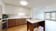 This 2-bedroom, 2-bathroom single-floor residence offers a thoughtfully designed layout that balances comfort and functionality. The kitchen is a standout feature, boasting two-tone cabinetry with white upper cabinets and warm wood-toned lower cabinets, white countertops, and a full suite of stainless steel appliances including a refrigerator, range, microwave, and dishwasher. A central kitchen island with a white surface provides additional prep space and casual seating. Light hardwood flooring runs throughout the main living areas, adding warmth and continuity. The living room is bright and open, featuring a large sliding window that invites natural light and offers outdoor views, complemented by track lighting and a baseboard heating system. The spacious primary bedroom features plush carpeting and a walk-in closet, while the second bedroom also offers carpeted flooring and a generous window with outdoor views. Both bathrooms are finished with warm wood vanities, white countertops, and hardwood flooring, with one bathroom featuring a shower-tub combination. A dedicated pantry and hallway add to the practicality of this well-organized residence.
