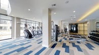 This single-floor fitness center is thoughtfully designed to support a comprehensive workout experience. The space is divided into distinct functional zones, offering both cardio and strength training areas to accommodate a wide range of exercise routines. The cardio section is lined with a row of stair climbers and elliptical machines positioned along expansive floor-to-ceiling windows, allowing natural light to flood the space while providing views of the surrounding cityscape. The strength training area features a multi-station cable machine and a selection of resistance equipment, catering to various fitness goals. A mirrored wall runs along one side of the space, enhancing the sense of openness and allowing users to monitor their form during workouts. A dedicated glass-enclosed room at the rear provides an additional private training or studio area. The flooring throughout features a striking blue, grey, and white patterned carpet tile design that adds visual interest and defines the different workout zones. Recessed lighting and ceiling fans ensure the environment remains bright, comfortable, and well-ventilated. The overall ambiance is clean, modern, and professional, making this fitness center a functional and inviting amenity.