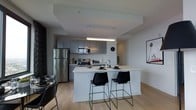 This single-floor space showcases a contemporary open-concept layout centered around a well-appointed kitchen. The kitchen features flat-panel cabinetry in a cool grey finish, paired with stainless steel appliances including a full-size refrigerator, microwave, and range. Light-colored countertops provide ample prep space and extend seamlessly into a central island that doubles as a breakfast bar, accommodating two black modern bar stools—an ideal setup for casual dining or entertaining. A sleek pendant light fixture hangs above the island, providing focused task lighting that complements the overall modern aesthetic. The light oak hardwood flooring flows throughout, adding warmth and continuity to the space. Neutral white walls and a clean ceiling design with subtle architectural detail contribute to the bright, airy atmosphere. A framed wall print adds a subtle decorative touch to the living area visible in the background. The thoughtful layout and cohesive design elements create a functional yet stylish environment well-suited for everyday living.