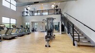 This two-story fitness center at 2000 Day Hill Rd in Windsor, Connecticut, is a well-appointed facility designed to support a comprehensive range of workout routines. The main floor features soaring double-height ceilings that lend a spacious, energizing atmosphere, accentuated by large black-framed windows that flood the space with natural light and offer views of the surrounding greenery. Light hardwood flooring runs throughout, complementing the clean white walls and industrial-style exposed ductwork overhead. The ground level is equipped with a full lineup of cardio machines, including multiple treadmills and stationary bikes, alongside a multi-station cable machine, free weights, weight racks, and adjustable benches — providing everything needed for a thorough strength and conditioning session. Floor-to-ceiling mirrors enhance the sense of openness and support form monitoring during workouts. A dedicated studio room off the main floor houses spin bikes, BOSU balls, and yoga mats, illuminated by natural light from multiple windows. A sleek black metal staircase leads to the open mezzanine level, where a studio space with mirrored walls, stability balls, resistance accessories, and a decorative wicker ceiling fan offers a quieter setting for stretching, yoga, or functional training, with scenic outdoor views visible from above.