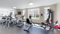 This fitness center offers a comprehensive workout environment spread across a single level, designed to accommodate a variety of exercise routines. The space is finished with durable rubber flooring throughout, crown molding along the ceilings, and bright white walls that contribute to a clean, welcoming atmosphere. Flush-mount ceiling lights provide consistent overhead illumination, while an abundance of large, multi-pane windows along multiple walls floods the space with natural light and offers views of the surrounding exterior grounds and building facades.

The cardio area is well-stocked with treadmills, elliptical trainers, upright and recumbent stationary bikes, and a rowing machine. A wall-mounted flat-screen television and a heart rate target chart are positioned for easy viewing during workouts. A dedicated strength and free-weight zone features a tiered dumbbell rack with a full range of weights, an adjustable workout bench, a ballet barre, and floor-to-ceiling mirrors that visually expand the space. A split-unit air conditioning system ensures climate control year-round. The entrance area includes safety features such as an emergency exit, fire extinguisher, and emergency phone.