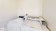 Located at 604 E Orange Grove Ave in Burbank, California, this dedicated laundry room is a compact, single-floor utility space designed to serve residents with practical, shared laundry amenities. The room features light-toned walls and neutral tile flooring that contribute to a clean, functional atmosphere. Natural light filters in through a white-framed window, brightening the interior and providing views of the surrounding neighborhood. The space is equipped with multiple coin-operated top-loading washing machines by Whirlpool, as well as a stacked washer and dryer unit, accommodating multiple users efficiently. A sturdy white utility door with dual ventilation grilles ensures proper airflow throughout the room, maintaining a comfortable environment during use. The straightforward, no-frills layout maximizes the usable area, keeping all machines accessible and the overall space easy to navigate. A notice board on the wall provides relevant usage information for residents. This well-lit, ventilated laundry facility offers a convenient and organized solution for everyday laundry needs.