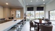Located at 14723 W Oaks Plaza Dr in Houston, TX, this single-story community clubhouse offers a thoughtfully designed space ideal for socializing and relaxation. The open-concept layout seamlessly connects a well-appointed kitchen and multiple lounge and dining areas, creating a welcoming and cohesive environment throughout.

The kitchen is a standout feature, showcasing a sweeping marble-look quartz countertop island with bar seating, gray shaker-style cabinetry, a decorative elongated tile backsplash, stainless steel appliances including a full-size refrigerator, and recessed lighting that enhances the clean, contemporary aesthetic.

Adjacent lounge and dining areas are furnished with round wooden tables and upholstered gray chairs, arranged to encourage conversation and gatherings. Large floor-to-ceiling windows flood the space with natural light and offer direct views of the outdoor pool area and surrounding landscaping, creating a bright and airy ambiance. Crown molding details add a refined architectural touch throughout.

A glass-partitioned section visible from the lounge opens to what appears to be a leasing or business center area, complete with a chandelier and curated artwork, adding an elegant focal point. Decorative shelving with stylish accents contributes to the modern, polished character of this well-designed community amenity space.