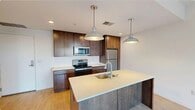 This contemporary single-floor unit offers a well-designed open-concept layout that balances functionality and modern aesthetics. The kitchen is a standout feature, showcasing rich dark-wood cabinetry paired with crisp white countertops, stainless steel appliances including an electric range, built-in microwave, and refrigerator. A center island with an undermount sink serves as the focal point, illuminated by a pair of stylish industrial-style pendant lights that add character to the space. Light hardwood flooring flows throughout the main living and kitchen areas, lending warmth and continuity. The living area is open and airy, bathed in natural light, with access to a private balcony through sliding glass doors that frame views of surrounding trees and the urban streetscape beyond. The bedroom is generously proportioned, featuring plush carpeting, large sliding glass doors that continue the outdoor connection, and ample wall space for flexible furniture arrangements. A secondary room with carpeting offers additional versatility. The bathroom is cleanly appointed with large-format tile flooring, a wide vanity with dark wood cabinetry and a white countertop, an oversized mirror, and a walk-in shower area. Clean white walls throughout the unit enhance the sense of brightness and space.