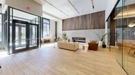 This thoughtfully designed single-floor space offers a generous, open layout that balances social gathering and quiet retreat. The entrance is defined by dramatic floor-to-ceiling glass walls and large glass doors, flooding the interior with abundant natural light and providing expansive views of the surrounding streetscape. Light wood-look flooring runs throughout, lending warmth and continuity across all areas.

The main lounge area is anchored by a striking fireplace set against a rich, dark wood-paneled accent wall with a concrete surround, creating a bold focal point. Comfortable seating arrangements, including plush sofas and accent chairs, invite relaxation in this airy, high-ceilinged space. Track lighting overhead supplements the natural light throughout the day.

A glass-enclosed dining and collaborative area features pendant globe lighting, a built-in shelving unit, and a long communal table with colorful seating — ideal for group gatherings or work sessions. Arched niches with floral wallpaper accents add an artistic, boutique touch to one corner of the space, complemented by counter-height seating along a terrazzo-topped bar ledge. Two hallways connect the primary areas to additional spaces, including what appears to be a mail room or package area, rounding out this well-appointed and design-forward communal environment.