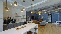 Located at 1817 Sir Tyler Dr in Wilmington, NC, this single-floor space makes a striking first impression with its thoughtfully curated interior design. The open-concept layout seamlessly connects the kitchenette and lounge areas, creating a cohesive and inviting environment throughout.

The kitchenette is outfitted with dark gray cabinetry, a clean white countertop island with bar seating, a built-in sink, and a beverage refrigerator. White subway tile backsplash adds texture, while a floating wood shelf provides functional display space. A bank of brass pendant lights with bell-shaped shades illuminates the prep area with warm, focused light.

The lounge area transitions naturally from the kitchenette and is anchored by rich navy blue walls adorned with decorative molding panels and gold-framed mirrors. A coffered ceiling with detailed trim adds architectural depth and sophistication to the space. Light wood flooring runs throughout, complementing the warm amber and cognac tones of the upholstered seating. A flat-screen television and an ornate floor mirror serve as standout focal points. The layered lighting scheme, combining recessed fixtures and a decorative pendant, enhances the space's warm and polished ambiance, making it well-suited for both productive and social gatherings.