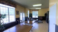 This single-story common area offers a welcoming and functional layout designed for gathering and relaxation. The room is finished with warm yellow walls accented by dark wainscoting along the lower portion, creating a two-tone effect that adds visual interest and depth. Tile flooring with a brick-like pattern runs throughout the space, providing a durable and easy-to-maintain surface.

Natural light enters generously through several windows fitted with patterned valances, as well as a set of white-framed French doors that lead to the outdoors, offering views of surrounding greenery. Overhead fluorescent lighting supplements the natural light, ensuring the space remains well-lit throughout the day.

The room is furnished with a comfortable seating arrangement, including a patterned sofa with accent pillows and an upholstered wingback chair positioned around a small coffee table atop an area rug. Dining or activity tables with accompanying chairs are placed toward the front of the room, supporting multiple uses. Decorative potted plants and framed wall art complete the space, contributing to a casual yet tidy communal atmosphere.