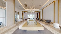 Located at 1710 N Congress Ave in West Palm Beach, Florida, this sophisticated single-floor lobby space presents a striking first impression through its contemporary design and upscale finishes. The interior spans a generously proportioned open floor plan that seamlessly blends multiple functional zones within one cohesive environment.

The architectural design is defined by soaring ceilings with layered coffered detailing and integrated cove lighting that casts a warm, ambient glow throughout. Gold-toned trim and framing elements accent the crisp white walls, creating an atmosphere of refined elegance. A stunning sculptural chandelier composed of globe-shaped glass pendants serves as a dramatic focal point, drawing the eye upward and anchoring the central space.

Flooring transitions thoughtfully between polished marble-look tile, geometric herringbone-patterned stone, and terrazzo, adding visual depth and texture underfoot. A central platform feature with a glass display element anchors the main reception area, flanked by a welcoming front desk with warm wood cabinetry and a white quartz counter.

Arched passageways with a warm plaster finish guide visitors through the space, leading toward floor-to-ceiling glass walls that flood the interior with natural light and offer direct views of a lush, palm-lined outdoor pool area. A comfortable lounge seating area with natural wood-framed furniture and marble wall panels provides an inviting retreat within the space.