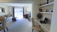 This single-floor residence offers a thoughtfully arranged layout that balances comfort and functionality. The living area is a welcoming space bathed in natural light, courtesy of large sliding glass doors that open to an outdoor patio, framed by elegant drape curtains. Plush carpeted flooring runs throughout, adding warmth underfoot, while a flat-screen television and cozy seating — including a rich burgundy armchair — make this an inviting space for relaxation. Decorative wall art adds a personal touch to the neutral-toned walls. Adjacent to the living area, a dedicated workspace is seamlessly integrated into the design, featuring a built-in floating desk and tiered open shelving above. The shelving provides ample display and storage space for books, personal keepsakes, and decorative objects. A patterned upholstered chair tucks neatly beneath the desk, completing this functional home office nook. The overall ambiance of the space is calm and residential, with a neutral color palette, soft lighting, and curated décor that together create a cozy, lived-in atmosphere suited to both productivity and leisure.